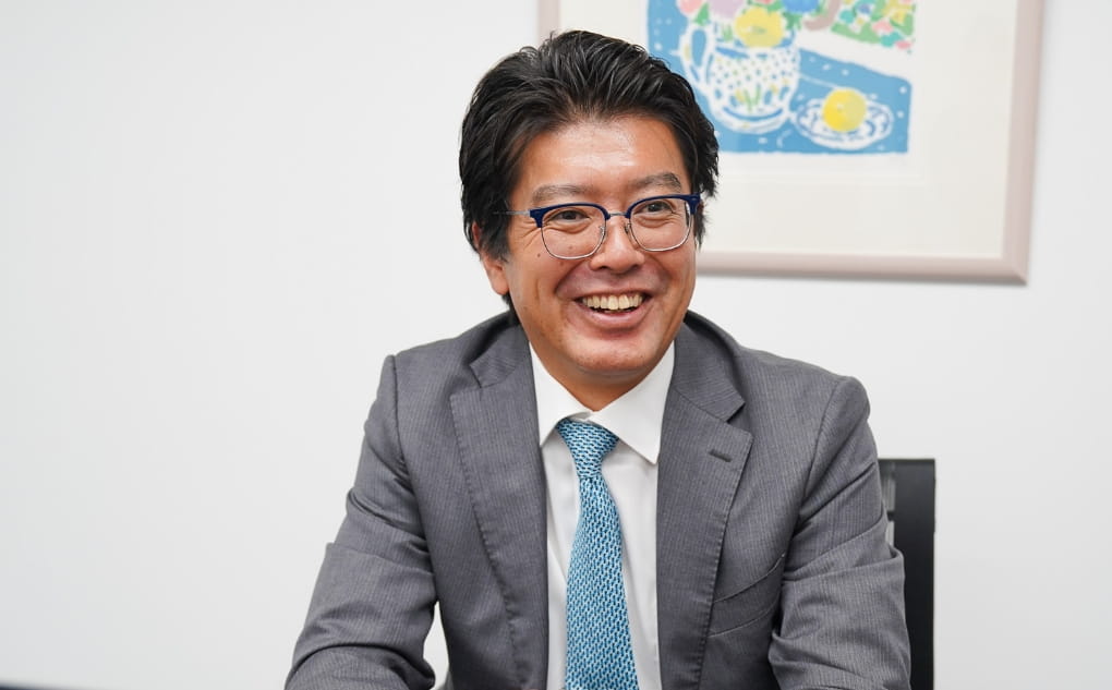 スーツを着た男性が笑顔でカメラに向かっている、穏やかで親しみやすい雰囲気が伝わる