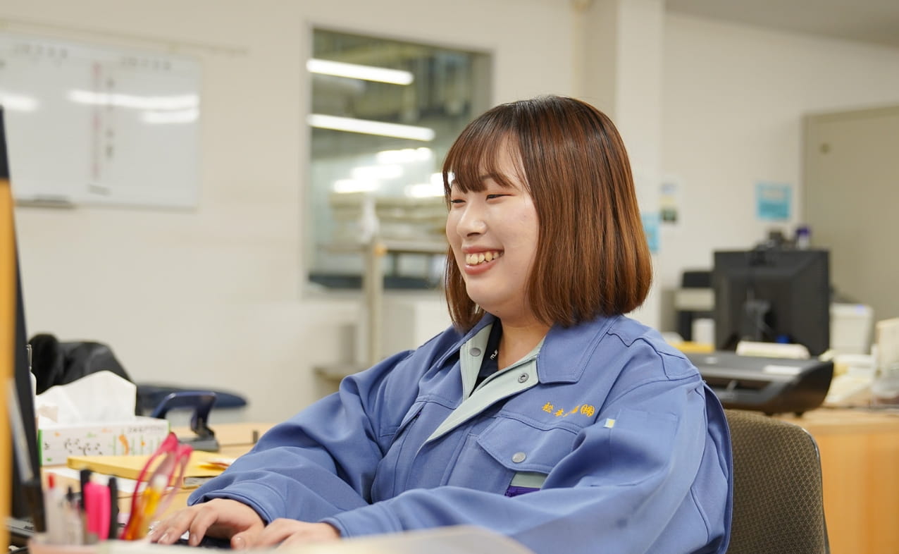 作業服を着た女性がデスクでパソコンを操作しながら笑顔を見せている。
