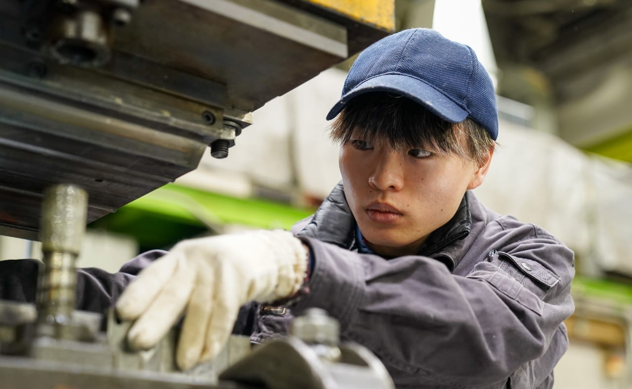 男性が工場内で機械の調整作業を真剣に行っている。