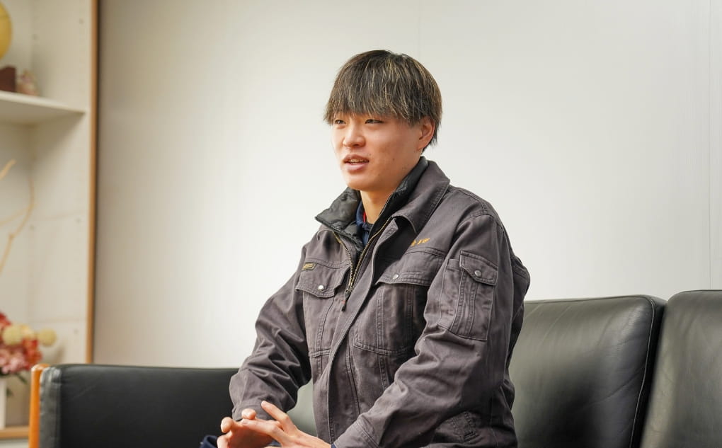 作業服を着た男性がソファに座り、真剣な表情で話をしている。