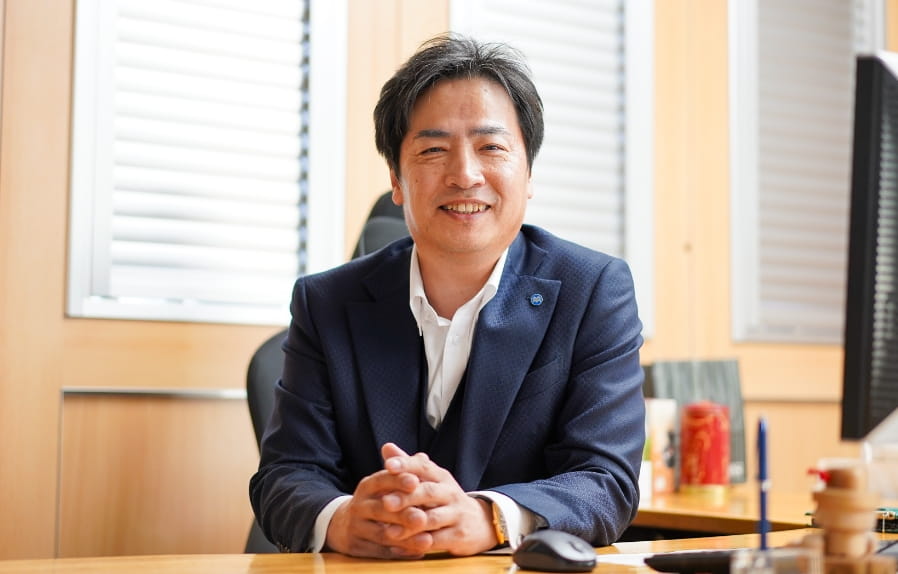松本金属株式会社の代表取締役がデスクに座り、笑顔でカメラに向かっている